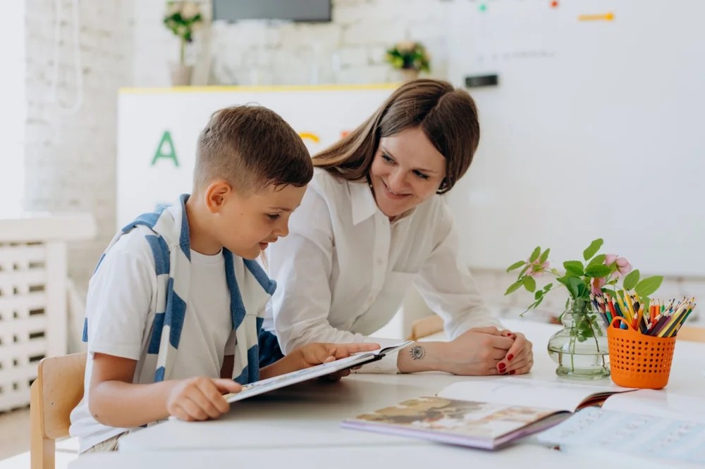 An educator supporting a young student at a classroom table.